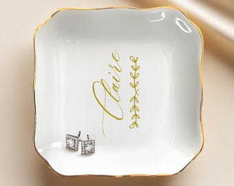 Può includere: Vassoio portagioie in ceramica bianca con bordo dorato e il nome "Claire" in oro. Un motivo a foglie dorate è accanto al nome. Due orecchini quadrati color argento con pietre trasparenti sono nel vassoio.