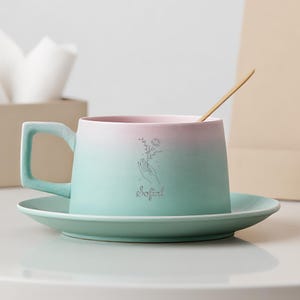 May include: A ceramic teacup and saucer set with a gradient color scheme of pink and teal. The cup features a handle and a delicate floral illustration with the word "Sofial". A wooden spoon rests inside the cup.