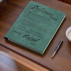 May include: A green journal with the word "Difference Maker" and definition printed on the cover. The journal also includes the name "Gabriel Gray" and a pen. The journal is on a wooden surface.