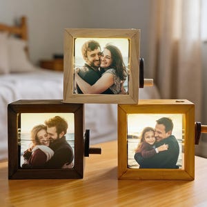 May include: Three wooden photo cubes displaying illuminated photographs of people. Each cube features a hand crank. The cubes are in shades of light, dark, and medium brown. The photos depict couples and a father with his daughter.