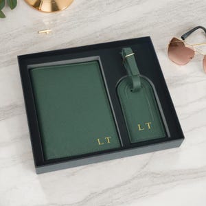 May include: A dark green travel set in a black box. The set includes a passport holder and a luggage tag, both with the gold initials "LT". The set is displayed on a marble surface.
