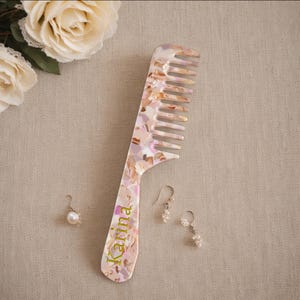 May include: A large, iridescent hair comb with a marbled pattern in shades of pink, beige, and gold. The name "Karina" is printed in green on the handle. Two pairs of pearl earrings are also present.