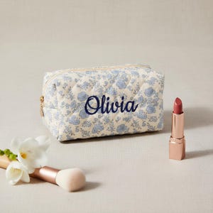 Puede incluir: Un neceser acolchado con estampado floral en crema y azul, bordado con el nombre "Olivia" en azul marino. Un labial y una brocha de maquillaje están al lado.