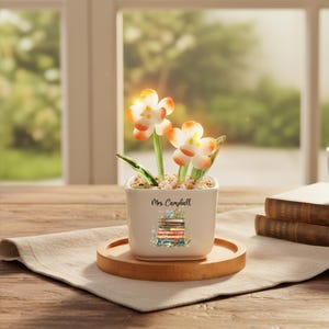 Può includere: Un piccolo vaso quadrato in ceramica bianca con un'illustrazione di una pila di libri e il nome "Mrs. Campbell" stampato. Il vaso contiene fiori decorativi con petali arancioni e bianchi. È posto su un vassoio di legno.