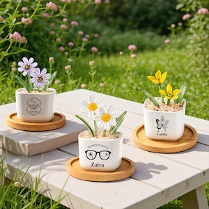 May include: Three white ceramic flower pots with personalized names and decorative flowers. Each pot sits on a round wooden coaster. The flowers include purple, white, and yellow designs. The pots have the names Lauren, Zaira, and Caitlin.