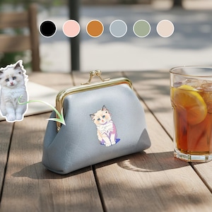 Pode incluir: Uma carteira azul clara com fecho dourado, com uma ilustração de um gatinho de desenho animado. A carteira está em cima de uma mesa de madeira, ao lado de um copo de chá gelado com limão. Um adesivo de gatinho também é visível.