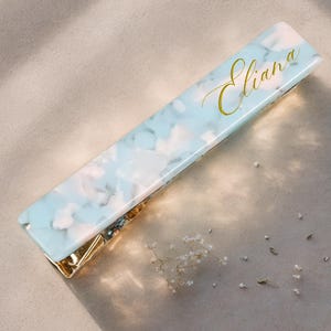 May include: A rectangular hair clip with a marbled blue and white design. The clip has gold-colored metal accents and the name "Eliana" in gold script. The clip is lying on a light-colored surface with small white flowers.