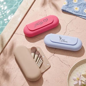 Puede incluir: Tres estuches de silicona para brochas de maquillaje en rosa, azul claro y beige. El estuche rosa tiene el nombre Hailey con un corazón, y el azul claro tiene el nombre Clana con un icono floral. El estuche beige está abierto, mostrando brochas.