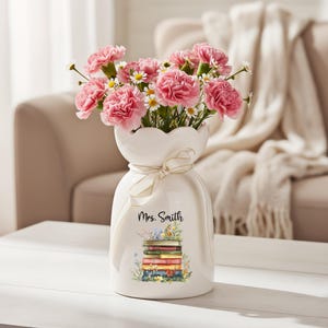 May include: A white ceramic vase with a scalloped top, tied with a cream ribbon, and filled with pink carnations and small white daisies. The vase has a watercolor design of books and the name "Mrs. Smith".