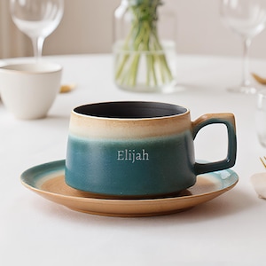 May include: A ceramic mug and saucer set with a two-tone design. The mug is teal and beige with a black interior and a handle. The saucer is teal and beige. The name "Elijah" is printed on the mug. The set is on a white surface.
