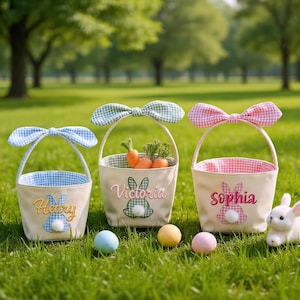 May include: Three Easter baskets with bunny ear handles and embroidered bunny designs. The baskets are beige with gingham lining and bows. One basket has "Henry" embroidered, another "Victoria", and the third "Sophia". Carrots and Easter eggs are in the grass.