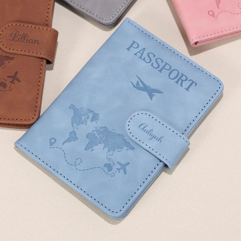 Op de afbeelding: Een lichtblauwe paspoorthouder met het woord "PASSPORT" en een wereldkaartontwerp. De houder heeft een flap met de naam "Aaliyah" en een gestippelde rand. Andere paspoorthouders in bruin, grijs en roze zijn zichtbaar.