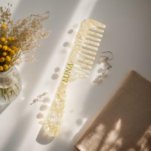 May include: A large, cream-colored comb with a marbled pattern and the word "LUNA" printed in gold. The comb is next to a pair of pearl earrings, a beige cloth, and a small vase of yellow flowers.