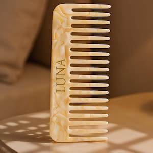 May include: A cream-coloured comb with a marbled pattern and the word "LUNA" in gold lettering. The comb has a wide-tooth design and is made of a smooth, glossy material. The comb is designed for hair care.