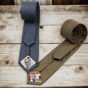 May include: Two neckties on a wooden surface. One is a blue-gray color with a diamond-shaped photo of a couple. The other is a rolled-up olive-green tie with a diamond-shaped photo of a family. Both ties have a textured appearance.