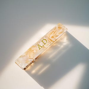 May include: Rectangular hair clip with a marbled cream and gold pattern. The clip features the gold letters "A.D." and a gold-colored metal clasp. The clip is angled on a white surface, with a shadow.