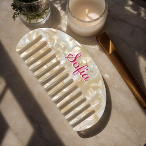 Puede incluir: Un peine blanco nacarado con mango curvo y dientes. El peine tiene el nombre "Sofia" en escritura rosa. También se ven un tubo de rímel dorado y una vela encendida.