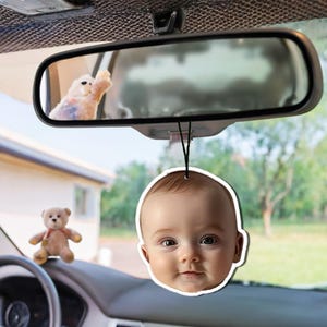 May include: A white car air freshener with a photo of a baby's face. The air freshener is hanging from the rearview mirror.
