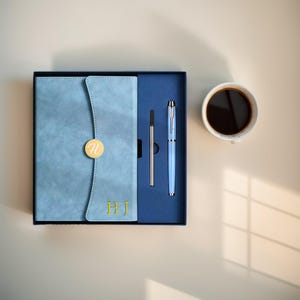 May include: A light blue notebook and pen set in a black box. The notebook has a flap with a gold-colored circular accent and the initials "HJ" in gold. A light blue pen and a pen refill are also included. A cup of coffee sits to the right.