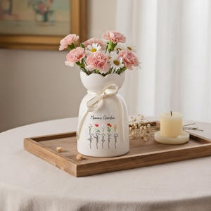 May include: A white ceramic vase with a scalloped top, tied with a cream ribbon, and filled with pink carnations and white daisies. The vase is inscribed with "Mama's Garden" and names. A lit candle sits nearby.