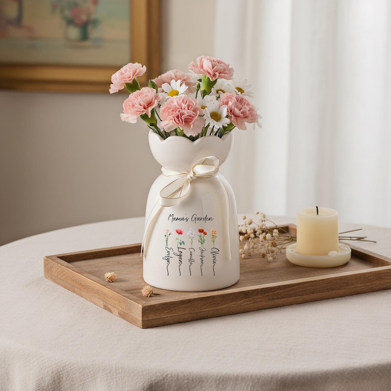 May include: A white ceramic vase with a scalloped top, tied with a cream ribbon, and filled with pink carnations and white daisies. The vase is inscribed with "Mama's Garden" and names. A lit candle sits nearby.