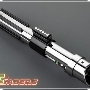 May include: A metallic silver lightsaber with black accents and a series of buttons. The lightsaber has a cylindrical design with a detailed handle and a glowing green and red light. The image includes the text "TSABERS" in a neon-style font.