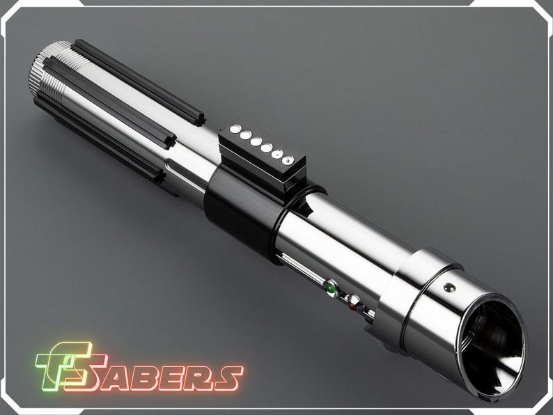 May include: A metallic silver lightsaber with black accents and a series of buttons. The lightsaber has a cylindrical design with a detailed handle and a glowing green and red light. The image includes the text "TSABERS" in a neon-style font.