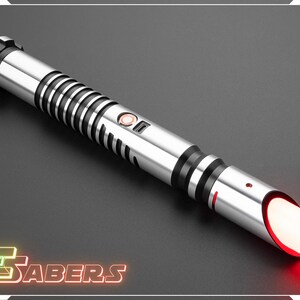 Puede incluir: Un sable de luz plateado y negro con una hoja roja brillante. El mango tiene anillos negros y un bot&oacute;n. El extremo del sable de luz tiene un brillo rojo. El logotipo "TSabers" est&aacute; en la esquina inferior izquierda.