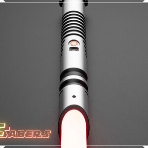 Puede incluir: Un sable de luz plateado con detalles negros y una hoja roja brillante. El mango presenta un dise&ntilde;o acanalado y un bot&oacute;n rojo. El logotipo "FSabers" est&aacute; en la esquina inferior izquierda.
