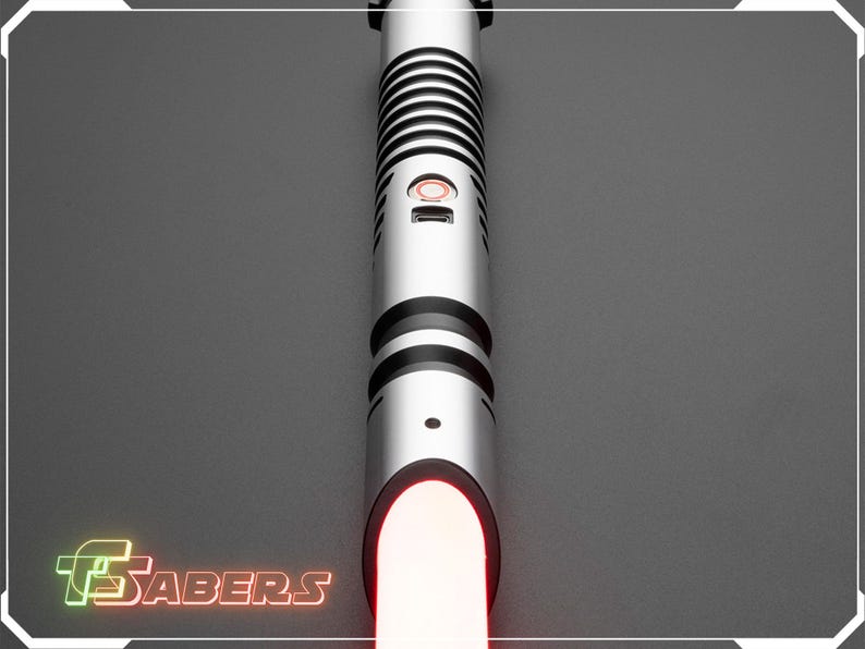 Puede incluir: Un sable de luz plateado con detalles negros y una hoja roja brillante. El mango presenta un dise&ntilde;o acanalado y un bot&oacute;n rojo. El logotipo "FSabers" est&aacute; en la esquina inferior izquierda.