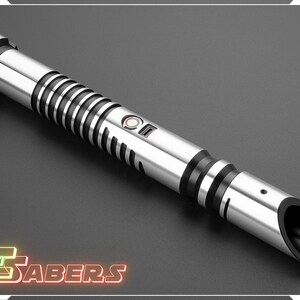 Puede incluir: Un sable de luz plateado y negro con un dise&ntilde;o cil&iacute;ndrico. El mango presenta anillos negros y un bot&oacute;n. La punta tiene una abertura negra. La imagen incluye el texto "TSABERS" en una fuente de estilo ne&oacute;n.