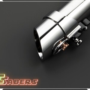 May include: Close-up of a chrome lightsaber hilt with intricate details. The hilt features a polished, reflective surface and a complex design with metallic accents. The image includes the text "TSABERS" in a neon-style font.