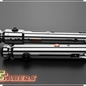 Puede incluir: Dos sables de luz cromados y pulidos se muestran sobre una superficie gris oscuro. Los sables de luz tienen componentes met&aacute;licos detallados, incluyendo botones y empu&ntilde;aduras cil&iacute;ndricas. El logotipo "TSABERS" est&aacute; en la parte inferior de la imagen.