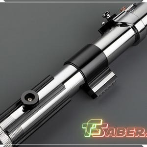 May include: A metallic silver and black lightsaber prop with intricate detailing. The prop features a cylindrical design with a black grip and various mechanical components. The image includes the text "TSABERS" in a neon-style font.