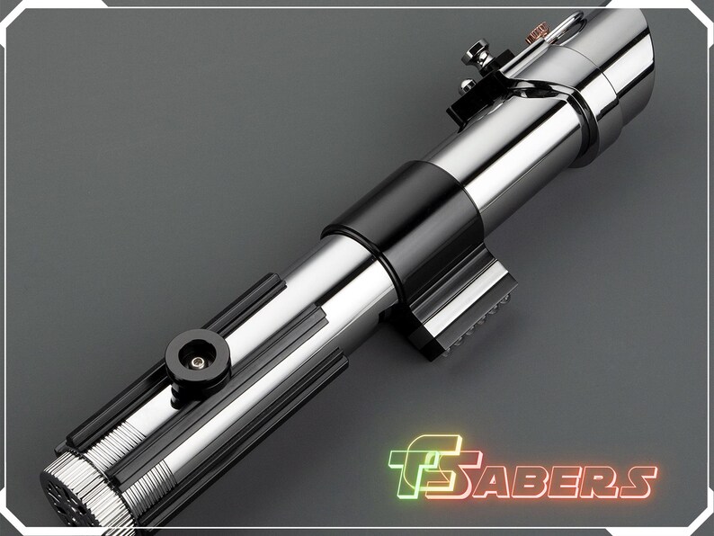 May include: A metallic silver and black lightsaber prop with intricate detailing. The prop features a cylindrical design with a black grip and various mechanical components. The image includes the text "TSABERS" in a neon-style font.