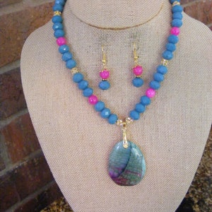 May include: A teal and pink beaded necklace with a gold pendant featuring a large, multi-colored agate stone. The necklace is displayed on a fabric-covered mannequin.