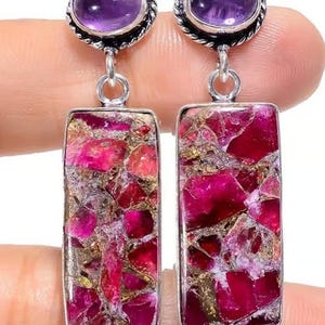 May include: A pair of dangle earrings featuring a purple amethyst stone at the top and a rectangular, multi-colored stone with shades of pink, red, and gold. The earrings are connected by a silver-colored metal. The earrings are held by a person's hand.