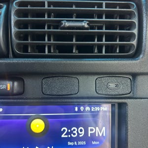 May include: Close-up of a car dashboard with a black air vent, buttons, and a digital display. The display shows the time, date, and various app icons. The time displayed is 2:39 PM.