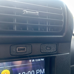 May include: Close-up of a car dashboard featuring a black air vent and two USB ports. The screen shows the time as 12:00 PM. The dashboard is black with a textured finish.