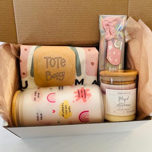May include: A gift box filled with a tote bag, a pink hair scrunchie, a candle, and a small jar. The tote bag has a pink and green pattern with the words "Tote Bag" on it. The candle is labeled "Masjid Al-Haram" and the jar has a rainbow design with the words "Keep it Halal" and "No worries, I've got uudu" on it.