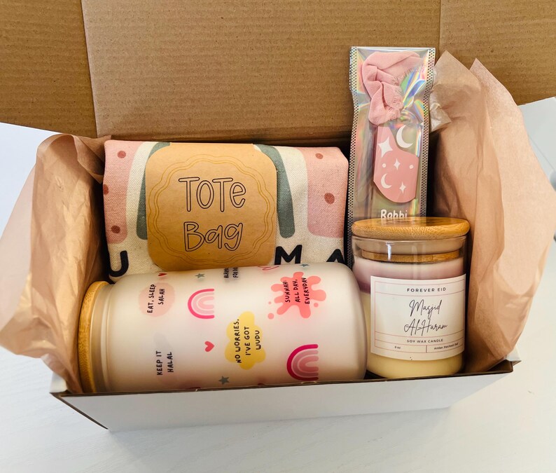 May include: A gift box filled with a tote bag, a pink hair scrunchie, a candle, and a small jar. The tote bag has a pink and green pattern with the words "Tote Bag" on it. The candle is labeled "Masjid Al-Haram" and the jar has a rainbow design with the words "Keep it Halal" and "No worries, I've got uudu" on it.