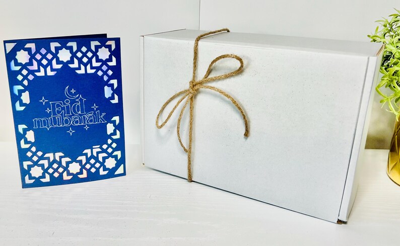 May include: A white gift box tied with twine and a blue card with a white geometric pattern and the text "Eid Mubarak".