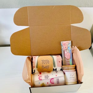 May include: A gift box filled with various items, including a tote bag with the text "Tote Bag", two mugs with rainbow designs and inspirational quotes, a candle with the text "Magical Afternoon", and a pink and blue hair tie.