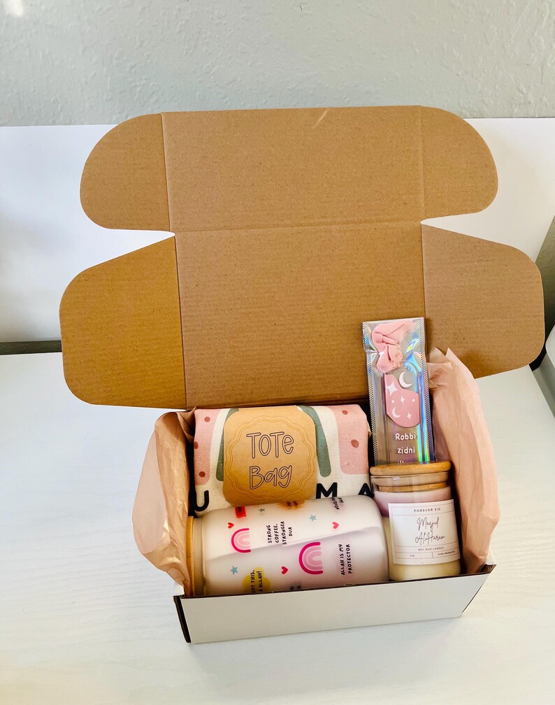 May include: A gift box filled with various items, including a tote bag with the text "Tote Bag", two mugs with rainbow designs and inspirational quotes, a candle with the text "Magical Afternoon", and a pink and blue hair tie.