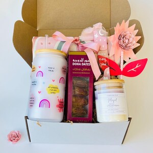 May include: A gift box filled with Ramadan themed items, including a pink and gray striped blanket, a white tumbler with rainbow designs and the text "Keep it Halal", a box of dates with the text "Doha Dates" and "Ramadan Mubarak" in Arabic, a white candle with a wooden lid and the text "Magel Alftar", and a pink and red paper flower.
