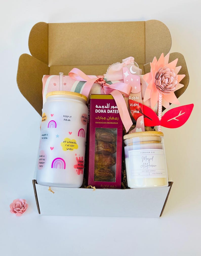May include: A gift box filled with Ramadan themed items, including a pink and gray striped blanket, a white tumbler with rainbow designs and the text "Keep it Halal", a box of dates with the text "Doha Dates" and "Ramadan Mubarak" in Arabic, a white candle with a wooden lid and the text "Magel Alftar", and a pink and red paper flower.