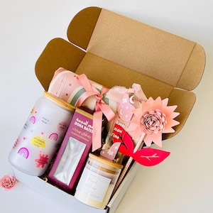 May include: A gift box filled with items for Ramadan. The box contains a pink and white blanket, a jar of dates, a candle, a glass jar with a pink and white design, and a pink flower with a wooden stick. The box is decorated with a pink ribbon and a tag that says "Robbi Zidni Ilma".