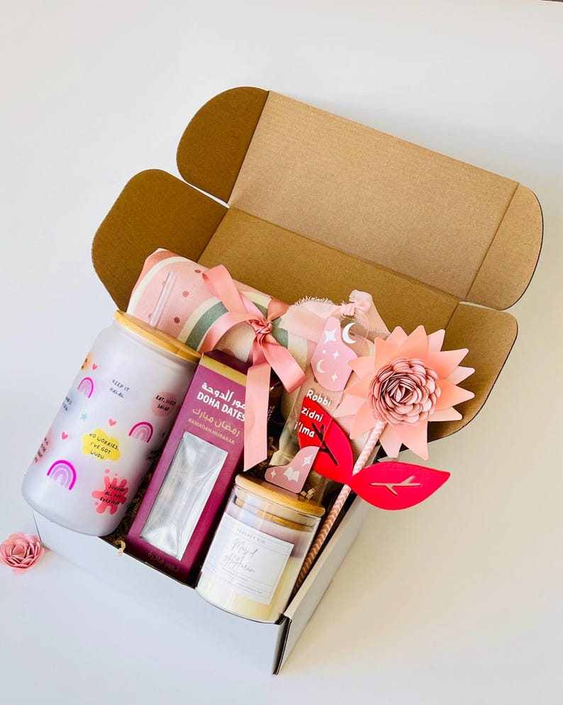 May include: A gift box filled with items for Ramadan. The box contains a pink and white blanket, a jar of dates, a candle, a glass jar with a pink and white design, and a pink flower with a wooden stick. The box is decorated with a pink ribbon and a tag that says "Robbi Zidni Ilma".
