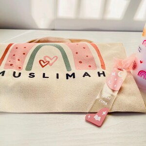 May include: A white canvas tote bag with a rainbow design and the word "MUSLIMAH" printed on it. The bag is accompanied by a pink and white glass jar with a wooden lid and a pink and white candle with the words "May Allah Bless You" printed on it.