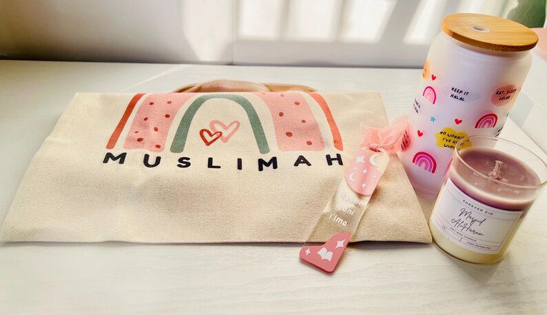 May include: A white canvas tote bag with a rainbow design and the word "MUSLIMAH" printed on it. The bag is accompanied by a pink and white glass jar with a wooden lid and a pink and white candle with the words "May Allah Bless You" printed on it.